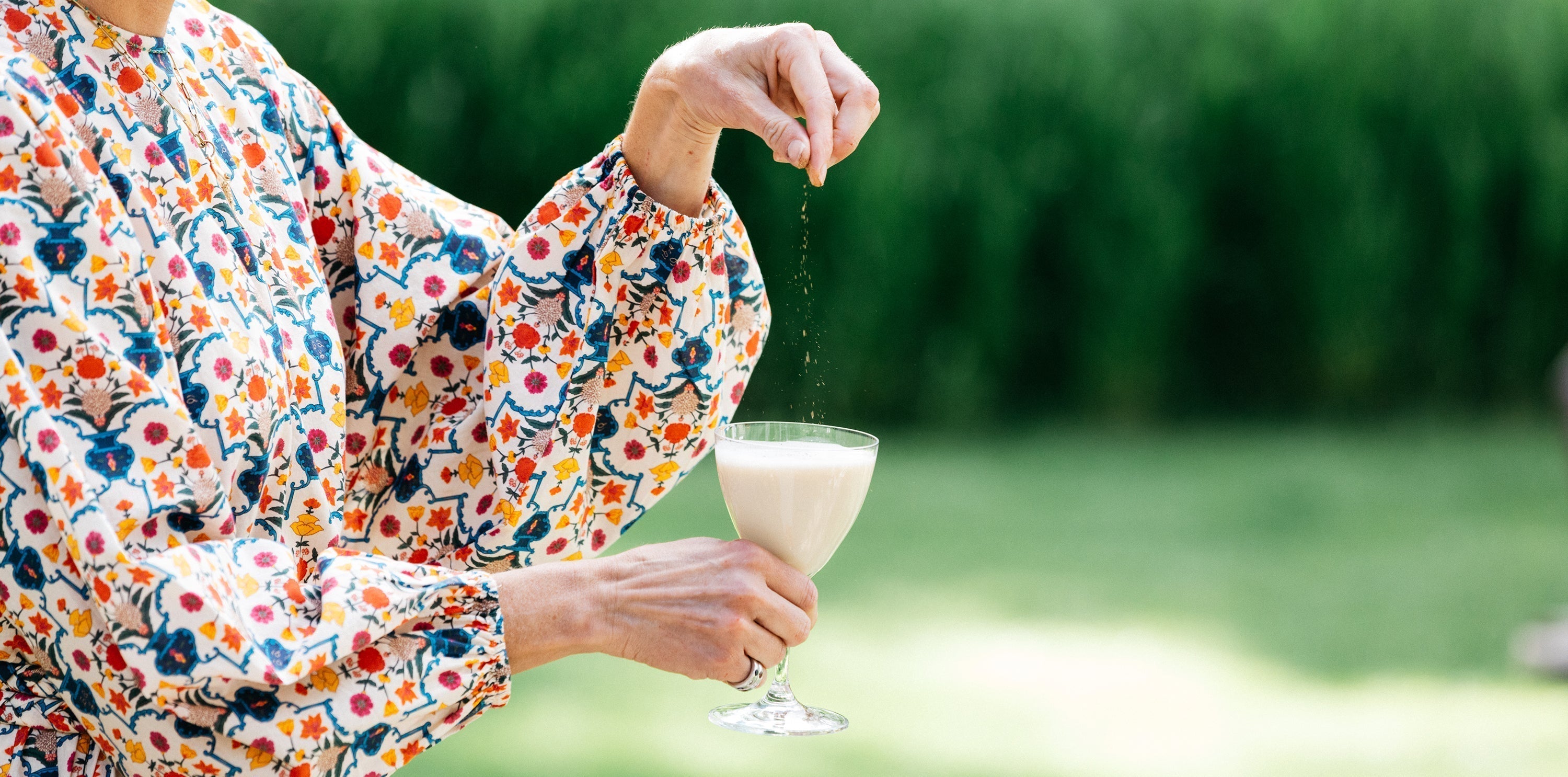 Woman in a floral blouse sprinkling spice onto a creamy drink outdoors for a refreshing summer beverage