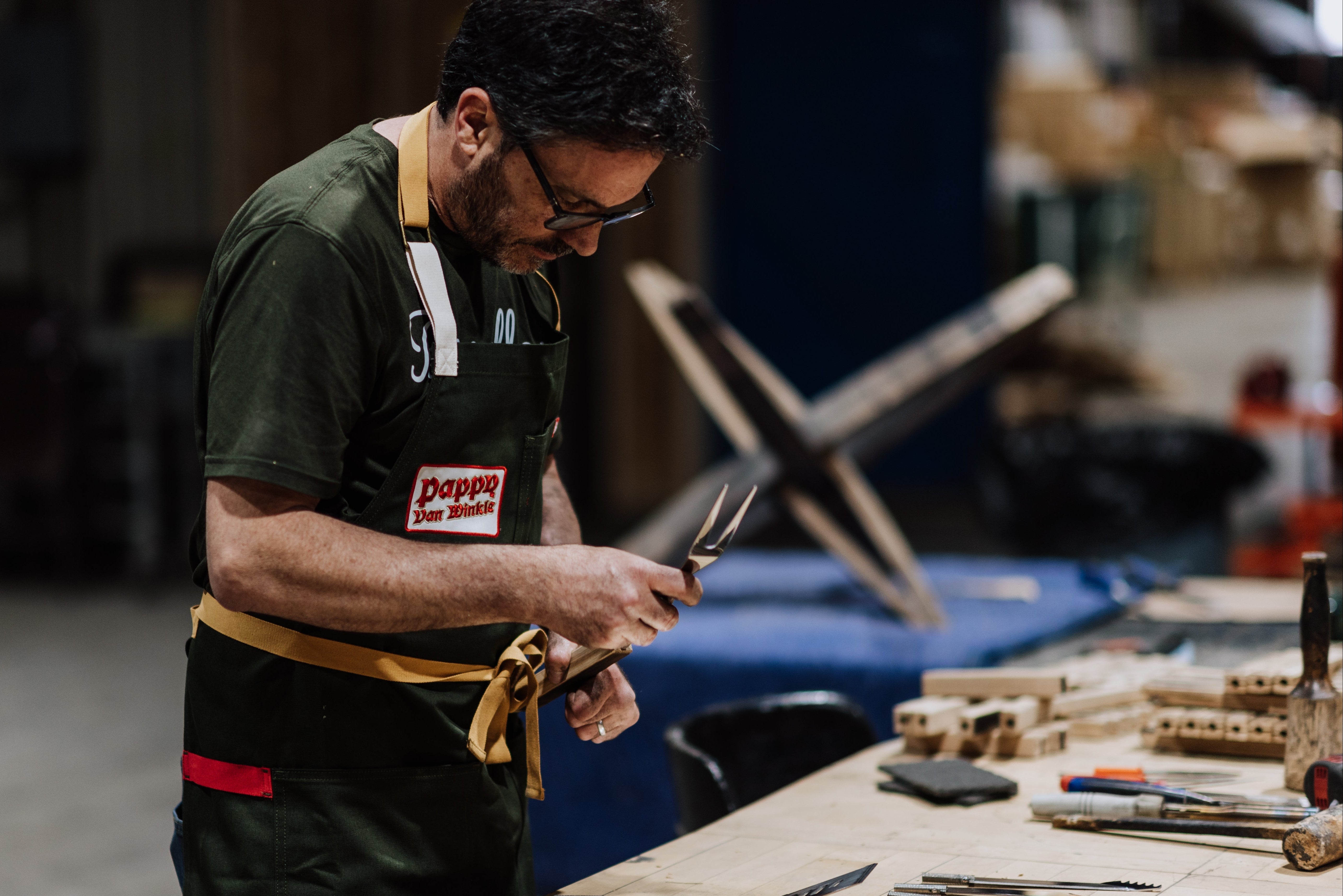 A man wearing a dark green apron with a 'Pappy Van Winkle' logo works at a wooden table filled with cut wood pieces and tools in a workshop setting.