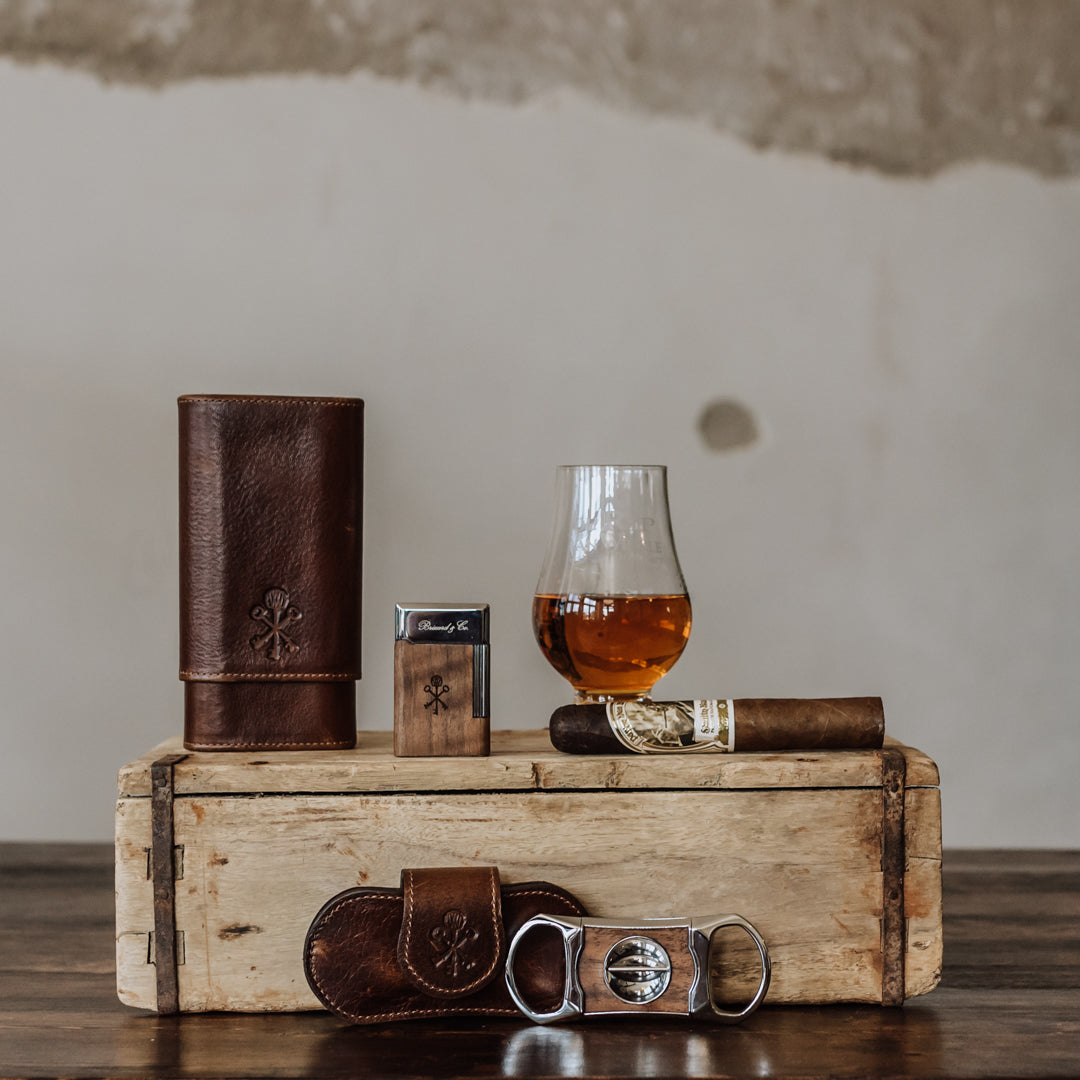 An Old Rip Van Winkle engraved Glencairn tasting glass on top of a wooden box with a cigar, lighter, cigar case, and two cigar cutters