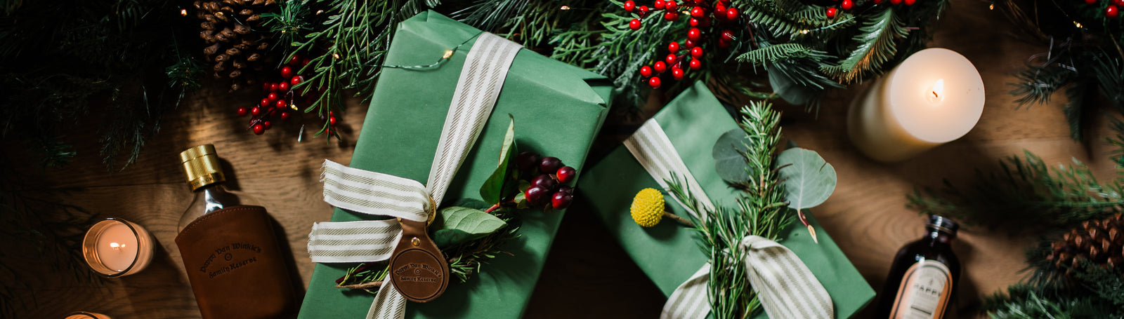 Two green Pappy & Company gift boxes with decorative ribbons on a wooden surface with candles and bottles.